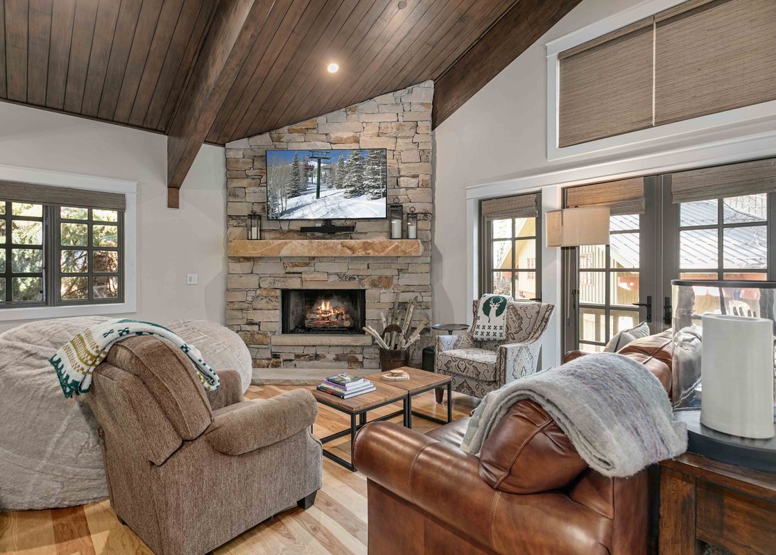 A cozy living room with a vaulted wooden ceiling, stone fireplace with a TV showing a snowy mountain scene, and comfortable seating with throws.