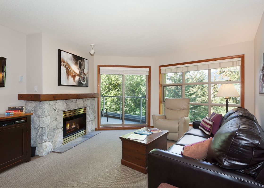 A cozy living room at Aspens on Blackcomb Unit 415 features a stone fireplace, comfortable leather sofa, and large windows offering views of lush greenery.