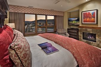 The primary bedroom at Cougar Mountain One Steamboat Place 501 features a fireplace, a king-sized bed, and large windows that offer a view of the surrounding mountains and village.