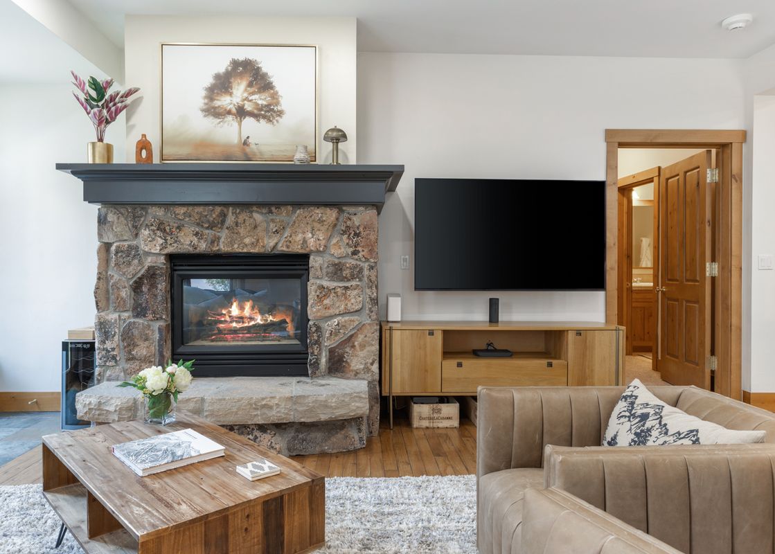 A cozy living room at 118 Villa Montane features a stone fireplace with a warm fire, a television mounted above a wooden media console, and comfortable seating.