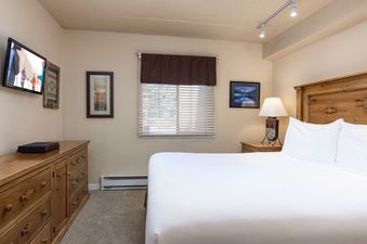 The master bedroom at Vantage Point 102 features a comfortable bed with white linens, a wall-mounted flat-screen TV above a rustic wooden dresser, and a window with blinds, creating a cozy and functional space.