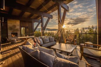 An expansive outdoor balcony at the Yeti Ridge Retreat offers comfortable seating areas, a dining table, and a fireplace, all set against a stunning backdrop of mountains and a cloud-filled valley during sunset.
