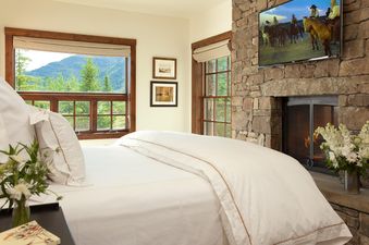 A cozy bedroom with a stone fireplace, flat-screen TV, and large windows looking out to mountains.