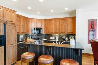 A well-equipped kitchen with wood cabinets, dark countertops, and a breakfast bar with three leather stools.