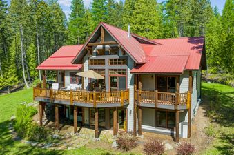 A large, multi-story lodge with a vibrant red roof and extensive wooden decks, surrounded by a dense forest on a sunny day.