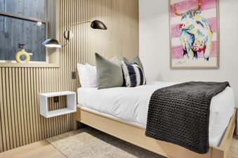 A serene bedroom at Woodside Manor features a bed with white and gray bedding, light wood paneling, elegant sconces, and a colorful highland cow painting on the wall.