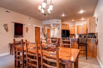 The image shows the dining area and an open-plan kitchen at Town Pointe B303, featuring a wooden dining table with rustic chairs, an elegant chandelier, and a breakfast bar with modern kitchen appliances.