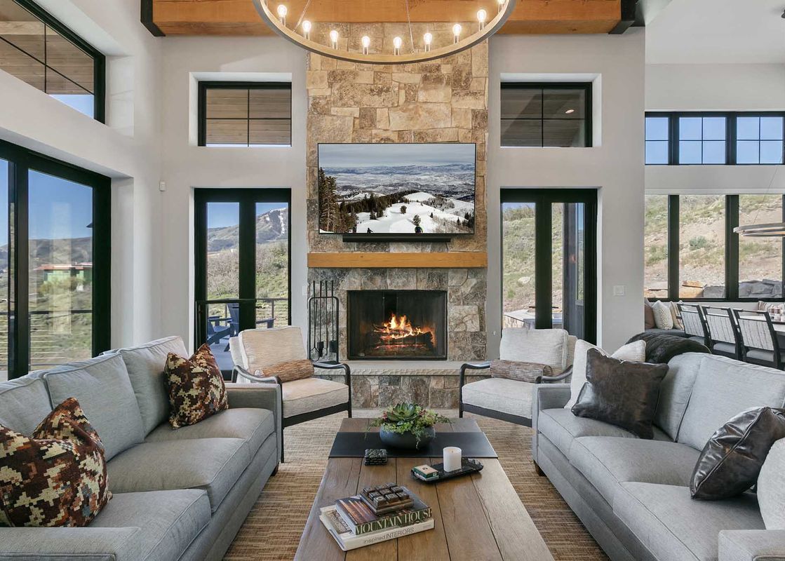 A spacious living room featuring a large stone fireplace with a roaring fire, a television displaying a winter mountain scene, two comfortable sofas with decorative pillows, a wooden coffee table, and expansive windows offering views of the mountains.