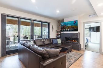 A luxurious living room in The Aerie features a large leather sectional sofa, a modern fireplace, a flat-screen TV, and sliding glass doors opening to a balcony with winter mountain views.