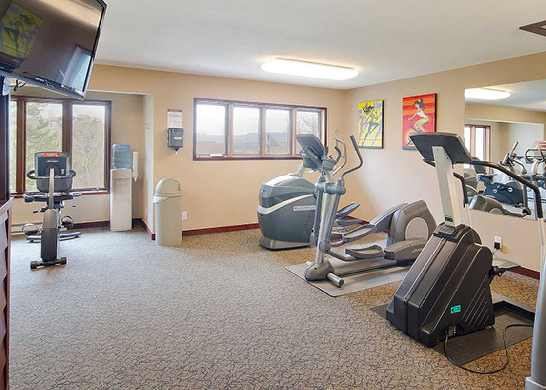 A well-equipped fitness center featuring various exercise machines, large windows, and a wall-mounted TV.