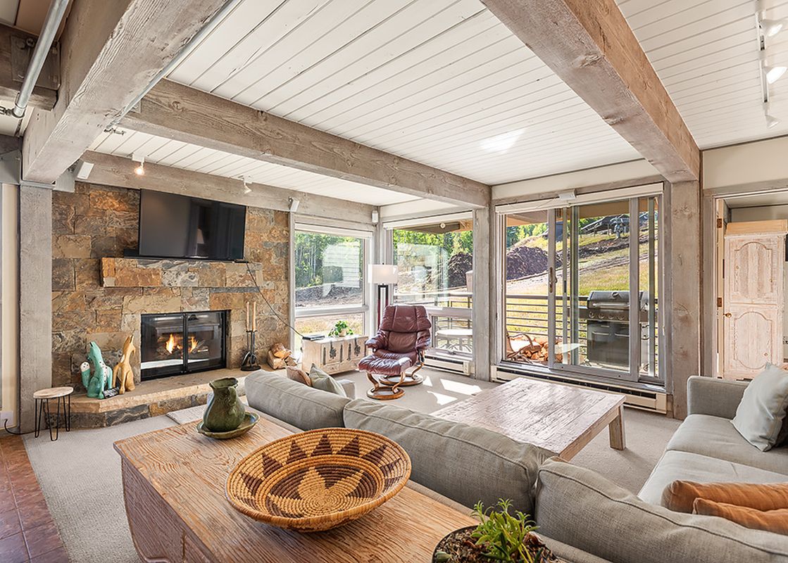 A spacious living room in Enclave 207 features a stone fireplace, a large gray sectional sofa, and a brown leather armchair, all under a ceiling with exposed wooden beams, with expansive windows looking out onto a patio and hillside.