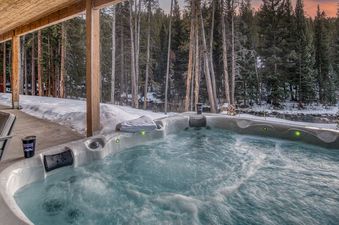 An inviting outdoor hot tub at the Everwild Chalet is set amidst a snow-covered forest with a creek, bathed in the soft glow of green lights, ready for a winter soak.