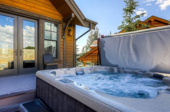 A private outdoor hot tub with bubbling water and a partially raised cover on the deck of Snowtop Manor, featuring wooden siding and French doors reflecting the sky.