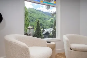 Two modern armchairs in a bright living room with a large window offering views of green mountains and houses.