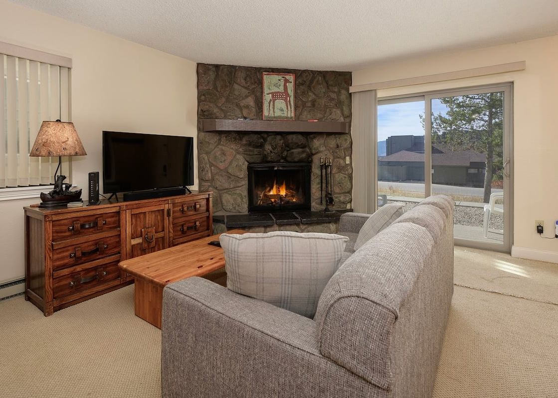 A cozy living room in Meadow Ridge 18 featuring a fireplace, a comfortable sofa, a wooden TV console with a flat-screen TV, and large sliding glass doors leading to an outdoor view.