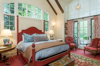 A red bed frame with a blue and white patterned duvet and pillows sits in a bedroom with a large window behind it, a rug with a red, green and brown pattern on the floor, and two chairs by a door that opens to a green view