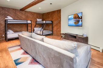 A bunk room at Pines Townhome 10 featuring two bunk beds, a sectional sofa, a wall-mounted TV, and a colorful area rug.