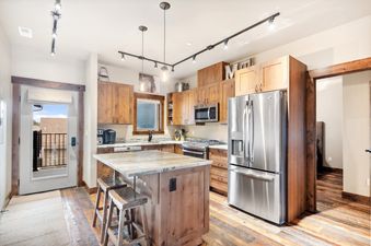 A modern kitchen in Baker Loft 303 featuring light wood cabinetry, stainless steel appliances, a marble-topped island with bar stools, and a door leading to a balcony.