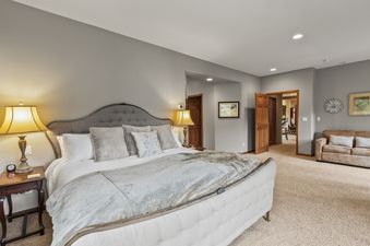 A spacious master bedroom with a large, tufted bed, soft lighting, and a cozy seating area, featuring neutral colors and elegant furnishings.