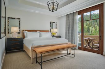 The bedroom of the Montage Four Bedroom Residence Resort View features a king-size bed with white linens, flanked by dark nightstands with lamps, a leather bench at the foot of the bed, and a private balcony overlooking a lush forest.