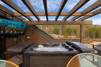 An outdoor hot tub with bubbling water sits on a balcony, overlooking a dense pine forest and distant mountains under a blue sky, with a television screen mounted nearby.