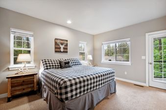 A cozy bedroom with a king-sized bed featuring a black and white buffalo check comforter, rustic wooden nightstands, and multiple windows offering views of lush trees.