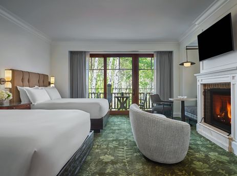 A spacious Montage Valley Queen Queen Room features two queen-sized beds, a cozy fireplace, and a balcony with an expansive view of lush trees.