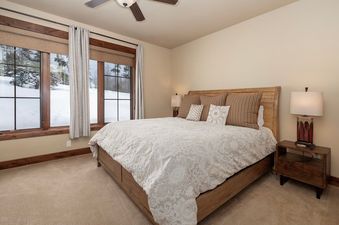 The primary bedroom at 751 Mountain Terrace features a large bed with a light-colored duvet and several pillows, along with a wooden headboard. A nightstand with a lamp is positioned to the right, and three large windows offer a view of a snowy landscape.