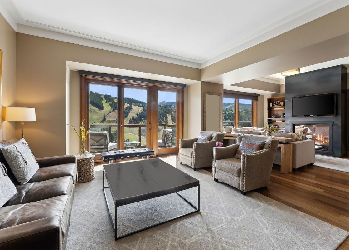 A spacious living room with a large window overlooking a mountain range, a leather sofa, two chairs, a coffee table, and a fireplace in the background.