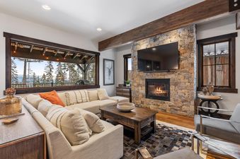 A luxurious living room at Inspiration Ski Haus featuring a large sectional sofa, a stone fireplace with a mounted TV, and a panoramic window overlooking a snowy forest.