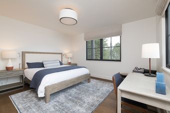 A bedroom with a bed, nightstand, and desk. There is a large window with a view of trees outside.