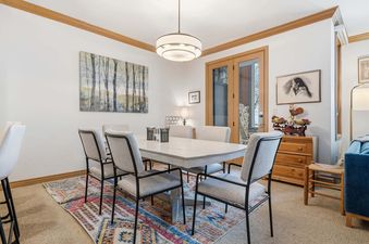 A rustic dining table with six chairs sits on a colorful rug in the dining area, illuminated by a modern chandelier, with a nature painting on the wall and French doors to the exterior.