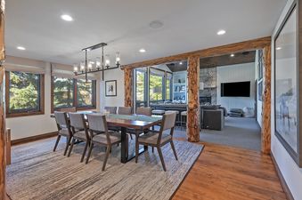 An expansive dining area and living room at the Antlers Estate, featuring a large wooden dining table with chairs, a modern chandelier, and access to a comfortable lounge space with a fireplace and television, creating a luxurious and inviting ambiance.