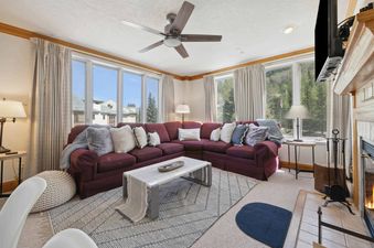 A cozy living room in The Charter L222 features a large burgundy sectional sofa with throw pillows, a coffee table on a geometric rug, and ample windows offering scenic views of trees and buildings.