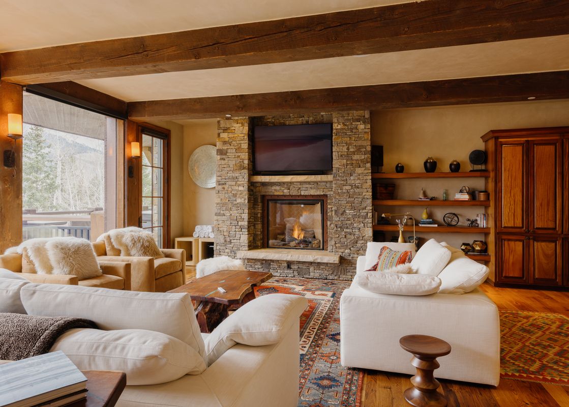 A cozy, rustic-chic living room with a fireplace, comfortable sofas, wooden beams, and a view of snowy mountains through large windows.
