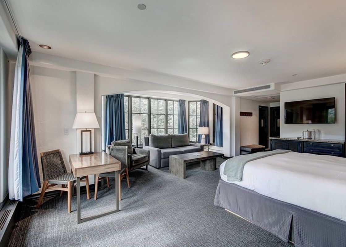 A spacious hotel room featuring a king-sized bed, a living area with a sofa and armchairs, a desk, and large windows with blue curtains.
