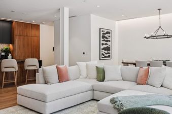 An expansive open-concept living area featuring a large, light-colored sectional sofa with decorative pillows, facing a modern kitchen with dark and wood cabinetry, and a dining area with a chandelier in the background.