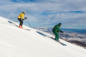 Two skiers descend a snowy mountain, with one in a yellow jacket and the other in a green suit, against a backdrop of distant mountains under a blue sky.