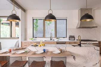 A modern kitchen with a large marble island and light wood cabinets. There are three pendant lights hanging above the island and a gold faucet at the sink.