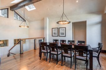 The dining area at 24 Little Cloud Trail, featuring a long dark wood table, eight chairs, two circular chandeliers, and a glass-paneled staircase.