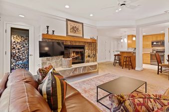 A cozy lounge area with a fireplace, TV, and brown leather sofa, with a modern kitchen visible in the background.