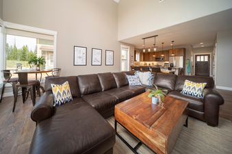 A spacious living room featuring a large brown leather sectional sofa, a wooden coffee table, and an open-concept kitchen area in the background.