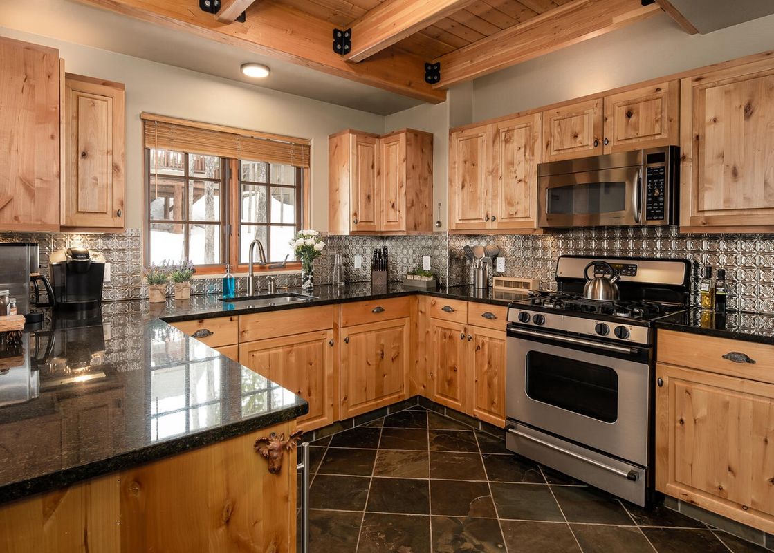 The kitchen of Moonlight Basin 5 Hidden Trail, showcasing wood cabinets and a stainless-steel stove and oven. The kitchen has a dark granite countertop with a large window that looks out onto the trees.