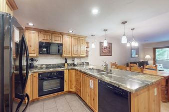 A modern kitchen with black appliances, granite countertops, light wood cabinets, and tile flooring, featuring a double basin sink and various kitchen essentials.