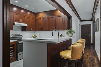 A modern kitchen with rich wood cabinetry, stainless steel appliances, light gray countertops, a sink with a gooseneck faucet, and an island bar with four yellow leather bar stools.