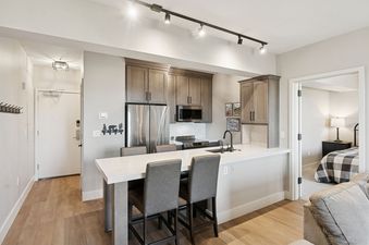 A modern kitchen and entryway in The Mason 3026, featuring sleek cabinetry, stainless steel appliances, an island with bar stools, and a glimpse into a cozy bedroom.