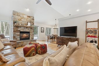 An inviting living room at the Meadow Lake Villa features a stone fireplace, comfortable leather sofas, and a dining area, all bathed in natural light from large windows showcasing a snowy outdoor view.