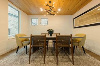 A modern dining room at Transfer Haus featuring a light wooden ceiling, a sleek dining table, and contemporary chairs, with large windows offering a view of the outdoors.