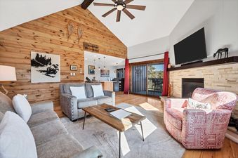 A bright and spacious living area features warm wooden paneling, comfortable seating, a stone fireplace with a mounted flat-screen TV, and sliding doors leading to a balcony.