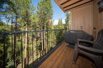 A private balcony at Beaver Village 1123 offers a Weber BBQ grill and a stunning view of a dense pine forest under a clear blue sky, with a hummingbird feeder hanging in the foreground.